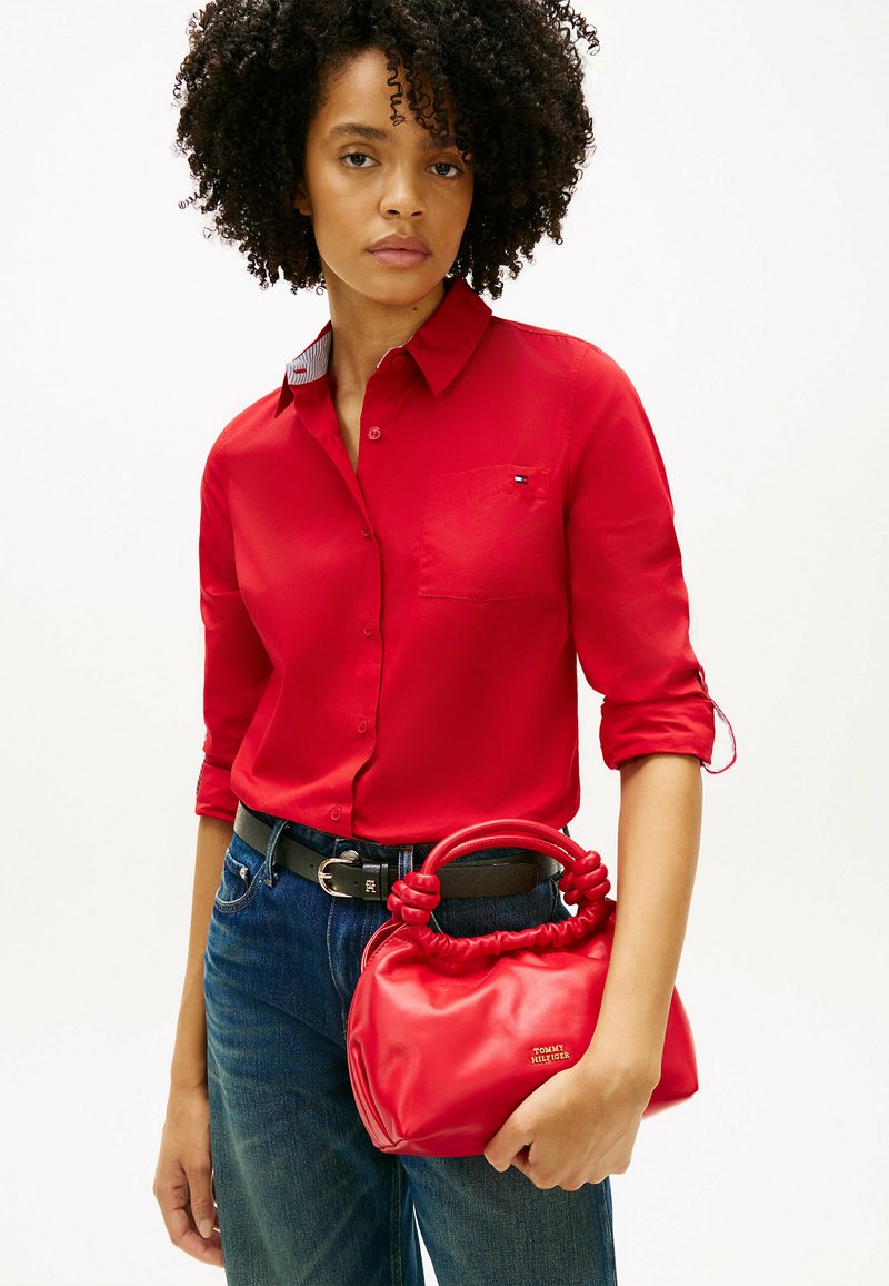 Femme aux cheveux bouclés portant une chemise rouge à boutons et un jean bleu, tenant un petit sac à main rouge avec des poignées nouées.