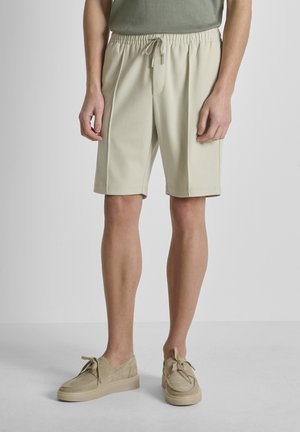 Uomo che indossa pantaloncini beige con coulisse e scarpe casual beige abbinate con lacci, in piedi contro uno sfondo chiaro uniforme.