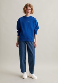 Blauwe sweatshirt met ronde halslijn en lange mouwen, gecombineerd met losse blauwe jeans en witte sneakers. Eenvoudige, casual outfit.