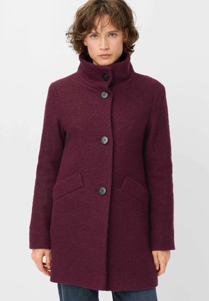 Woman wearing a dark purple wool coat with a high collar and four black buttons, standing against a plain light background.