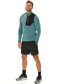 Langärmliges türkisfarbenes Sportshirt mit einer schwarzen Brusttasche, kombiniert mit schwarzen Shorts und grauen Laufschuhen, mit einem Viertel-Reißverschluss-Design.