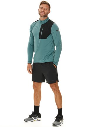 Langärmliges türkisfarbenes Sportshirt mit einer schwarzen Brusttasche, kombiniert mit schwarzen Shorts und grauen Laufschuhen, mit einem Viertel-Reißverschluss-Design.