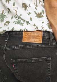 Black denim jeans with visible stitching, featuring a leather patch with branding. The shirt has a white background with green plant patterns.