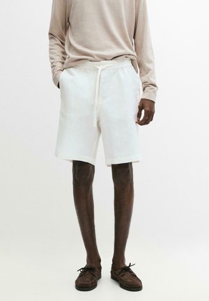 Uomo che indossa una camicia a maniche lunghe beige chiaro, pantaloncini bianchi con coulisse e mocassini marroni, in piedi davanti a uno sfondo bianco uniforme.