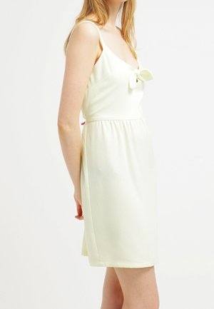 Vrouw die een mouwloze, crèmekleurige jurk draagt met een strikdetail aan de voorkant, staand tegen een effen witte achtergrond.