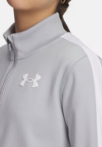 Svetlo šedá športová bunda s vysokým golierom, bielymi bočnými panelmi a bielym logom Under Armour na hrudi. Hladká textúra.