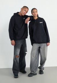 Svarta luvtröjor med framfickor, med vita logotyper. Modellerna bär slitna mörkgrå jeans och svarta sneakers, stående mot en ljus bakgrund.