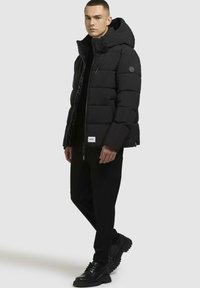 Schwarze Steppjacke mit Kapuze, mit gesteppten Abschnitten, einem Reißverschluss vorne und Logodetails. Getragen mit schwarzen Hosen und Stiefeln.