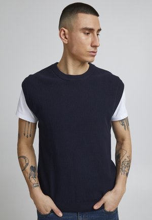 Suéter sin mangas de punto en azul marino con textura acanalada, superpuesto sobre una camiseta blanca. Presenta un cuello redondo y un ajuste relajado.
