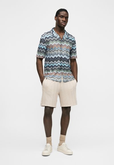 Άνδρας που φοράει κοντομάνικο πουκάμισο με πολύχρωμο ζιγκ ζαγκ μοτίβο, μπεζ σορτς, μπεζ κάλτσες και λευκά αθλητικά παπούτσια, στέκεται με τα χέρια στις τσέπες.