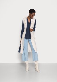 Colete bege sem mangas sobre uma blusa azul-marinho, combinado com jeans de perna larga azul-claro e botas de salto bege. Os acessórios incluem uma mala azul-marinho.