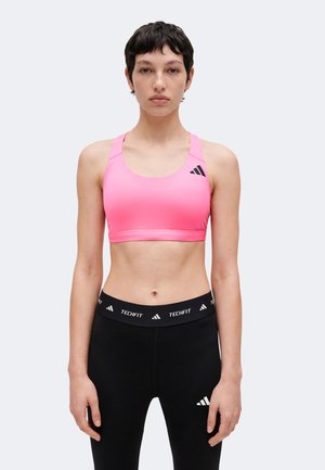 Femeie purtând un sutien sport roz și colanți negri cu sigle Adidas, stând în fața unui fundal simplu, deschis, cu fața spre înainte.