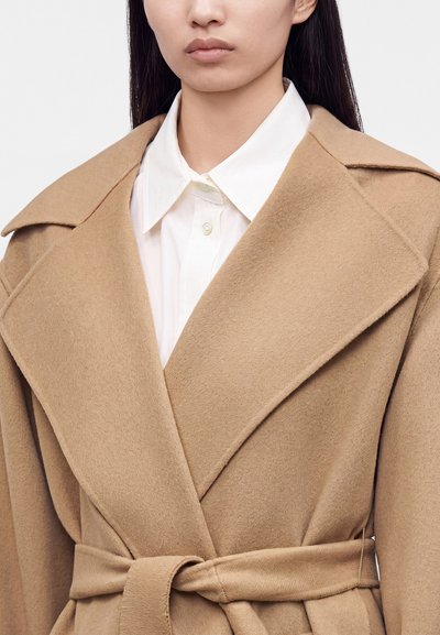 Femme portant un manteau en laine couleur camel ceinturé sur une chemise blanche à col, vue de la poitrine vers le haut sur un fond uni.