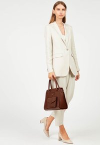 Costume beige clair sur mesure avec un blazer ajusté et un pantalon large, associé à un sac à main structuré marron et des chaussures à talons beiges.