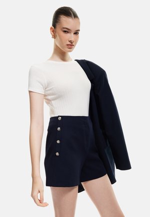 Weißes, geripptes Kurzarmoberteil kombiniert mit dunkelblauen, hochtaillierten Shorts, die silberne Knopfdetails an der Seite aufweisen.