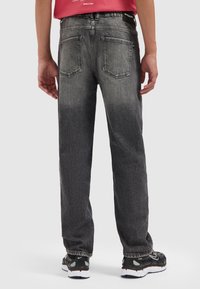 Person, der schwarze und grau ausgewaschene Jeans, ein rotes Shirt und schwarze Sneakers trägt, steht mit entspannten Armen an den Seiten vor einem weißen Hintergrund.