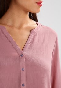 Blouse rose texturée avec un col en V et de petits boutons violets. Le tissu semble léger et lisse, adapté à une tenue décontractée.