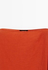 Roest-oranje rechthoekige textiel met een zichtbare rechte naad en een klein zwart label gecentreerd langs de bovenrand.