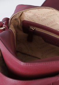 Interno della borsa in pelle bordeaux con una tasca con zip e foderato in tessuto con stampa logo ripetuta. Fibbia della cerniera in tono oro visibile.