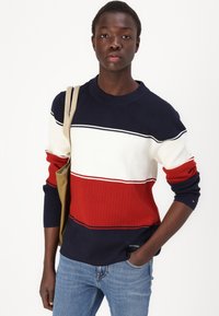 Pull en tricot avec des rayures horizontales bleu marine, blanches et rouges ; texture côtelée et encolure ronde ; présenté avec un jeans en denim et un sac beige.