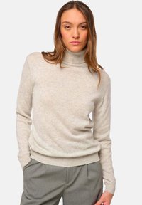 Pull à col roulé beige clair en tissu texturé doux, avec des poignets et un ourlet côtelés, porté avec un pantalon tailleur gris.