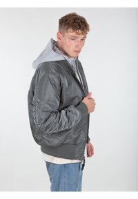 Giacca bomber grigia in satin con polsini e colletto a costine. Indossata sopra una felpa grigia chiara. Vista laterale, che mostra il design elegante e la texture liscia.