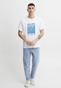 Camiseta de algodón blanca con un gráfico de agua azul y texto negro en la parte frontal, combinada con jeans de mezclilla azul claro y zapatillas blancas.