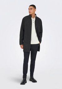 Mørkegrå kåpe med høy krage og knappestolpe foran, båret over en hvit skjorte. Slanke, svarte bukser og svarte støvler fullfører antrekket.