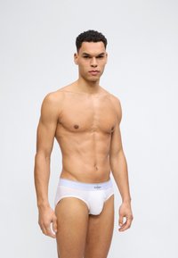 Καλοσχηματισμένος άνδρας με κοντά σγουρά μαλλιά, φορώντας λευκά εσώρουχα Calvin Klein, στέκεται μπροστά σε ένα απλό, ανοιχτό γκρι φόντο με ουδέτερη έκφραση.