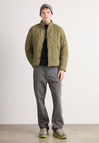 Joven vestido con una chaqueta acolchada verde oliva, gorro gris, camiseta negra, jeans grises holgados y zapatillas gris-verdosas, de pie contra una pared blanca.