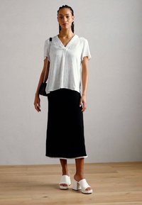 Blusa bianca a maniche corte con un motivo sottile, abbinata a una gonna nera fino al ginocchio. Indossata con sandali bianchi con tacco blocco. Borsa a tracolla nera.