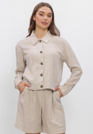 Jonge vrouw die een beige blouse met knopen en lange mouwen draagt en bijpassende losse shorts, staand met handen in de zakken tegen een effen achtergrond.