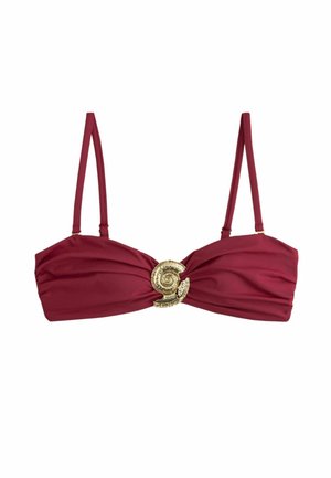 Frau trägt dunkelroten Bikini mit Muschel-Akzenten und gehäkeltem beigen Hut, steht am Wasser mit Armbändern am Handgelenk.
