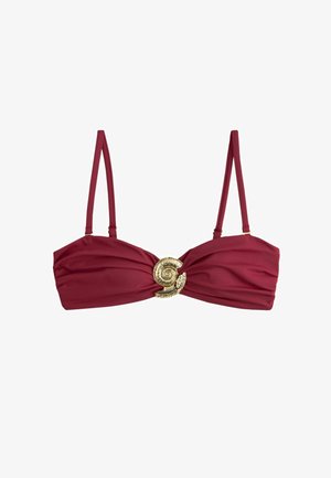 Kvinde iført mørkerødt bikini med skaller og hæklet beige hat står ved vandet med armbånd om håndleddet.