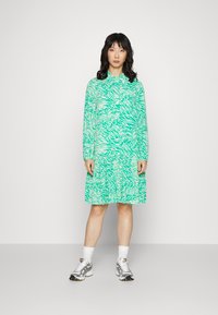 Vestido verde estampado com mangas longas, apresentando um design de riscas de zebra. O vestido tem uma bainha em camadas e é combinado com ténis prateados.