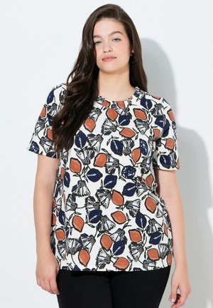 Ung kvinna med långt mörkt hår som bär en vit t-shirt med korta ärmar och blått och orange blommönster, stående mot en vit bakgrund.