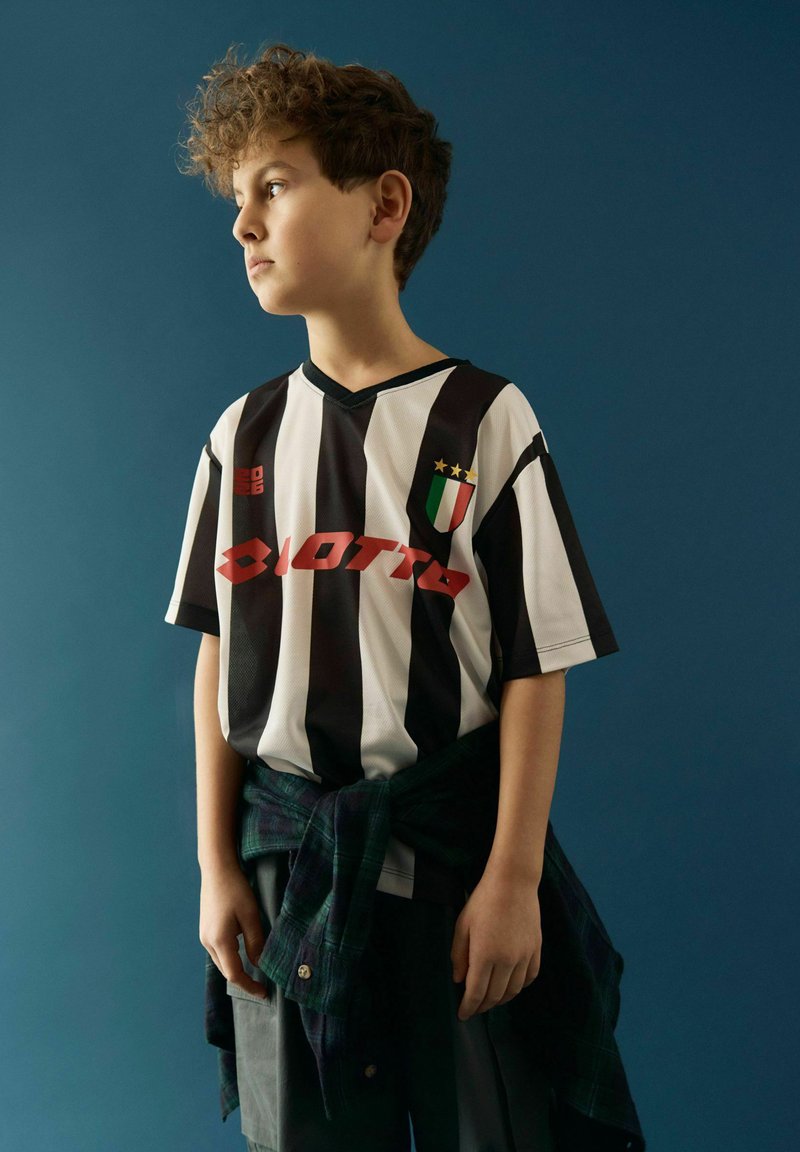 Garçon aux cheveux bouclés portant un maillot de football rayé noir et blanc et une chemise à carreaux foncés nouée autour de la taille, faisant face à gauche.