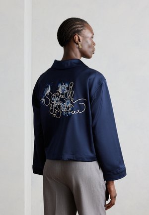 Frau mit kurzen geflochtenen Haaren, die eine marineblaue Jacke mit blauer Blumenstickerei und kursivem Text auf dem Rücken trägt, kombiniert mit hellgrauen Hosen.