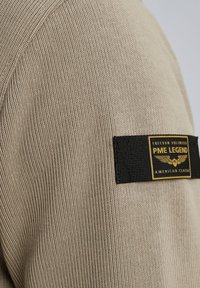 Tissu beige côtelé avec une étiquette rectangulaire noire et dorée indiquant PME Legend American Classic sur la partie supérieure de la manche.