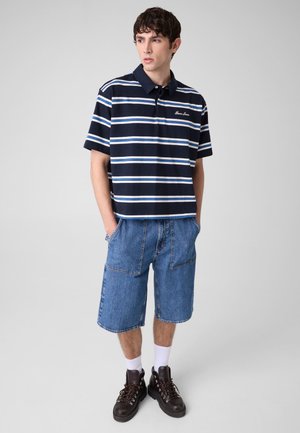 Jeune homme portant un polo rayé bleu marine, un short en jean, des chaussettes blanches et des chaussures de randonnée marron foncé, debout les mains dans les poches.