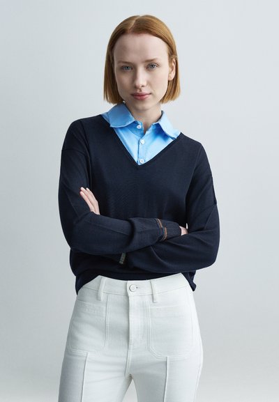 Pull marin en v superposé sur une chemise à col bleu clair, avec des accents arc-en-ciel sur les manches et un pantalon blanc taille haute.