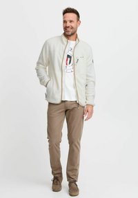 Veste en polaire beige légère avec fermeture éclair frontale, poches latérales, finitions de fermeture éclair contrastantes, portée sur un T-shirt graphique blanc et un pantalon marron.