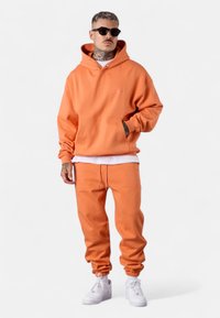 Orangener Hoodie mit einer vorderen Tasche und Zugband, kombiniert mit passenden Jogginghosen. Abgerundet mit weißen Sneakers, vor einem neutralen Hintergrund.