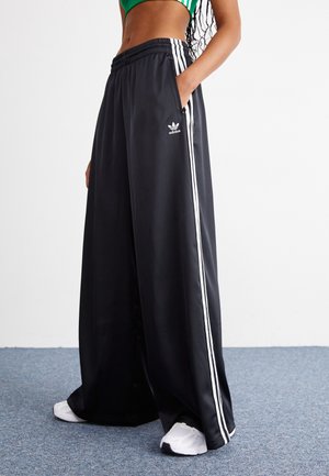 Osoba ubraną w czarne szerokie spodnie sportowe Adidas z białymi paskami i białe sportowe buty, stojąca na niebieskim wykładzinowym podłożu.