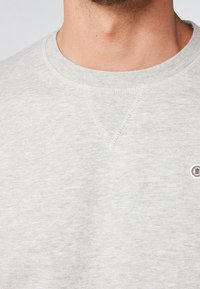 Sudadera gris clara hecha de una tela suave, con un cuello redondo, textura sutil y un pequeño logo circular.