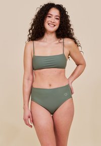 Kobieta z kręconymi włosami uśmiechająca się, ubrana w oliwkowo-zielony bikini dwuczęściowy na jednolitym beżowym tle.