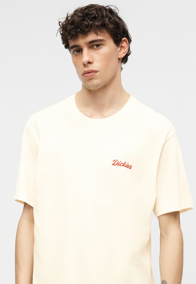 Joven con cabello rizado, vistiendo una camiseta Dickies color crema y un pequeño pendiente de aro en un fondo gris claro.