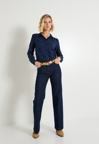 Blusa texturizada azul marino con botones y cuello, combinada con pantalones de mezclilla oscuros de cintura alta. Cinturón dorado, botas de tacón marrones.