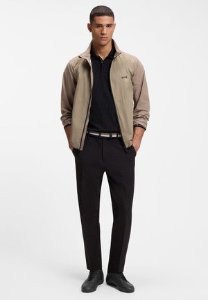 Jonge man die een beige jas met ritssluiting draagt over een zwart shirt, zwarte broek, zwarte schoenen en een gespenriem met patroon, staand met handen in de zakken.