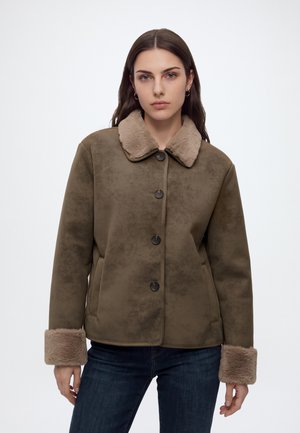 Femme aux longs cheveux bruns porte une veste boutonnée vert olive avec col et poignets en fausse fourrure beige, associée à un jean bleu foncé.
